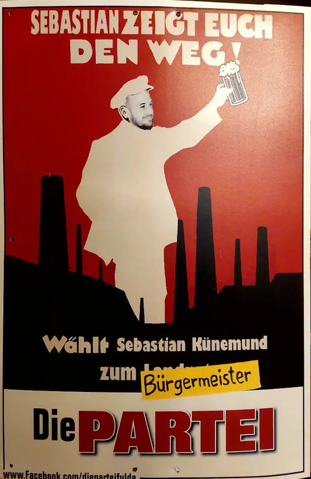 Ein Wahlplakat zeigt Sebastian Künemund als weiße Silhouette vor einem durch schwarze Schornsteine geprägtem Gebiet. Mit dem linken Arm streckt er ein Glas Bier in die Höhe.